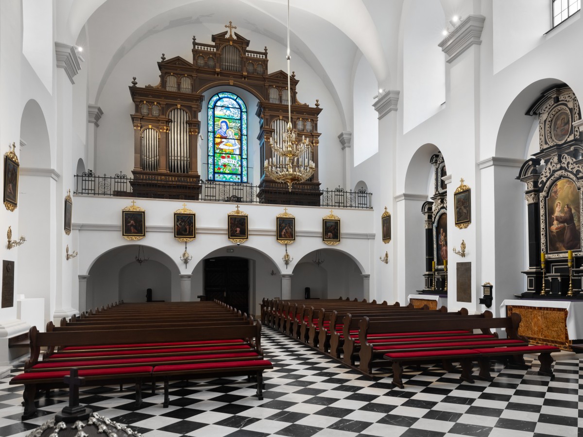  Ein barockes Kirchenschiff mit Reihen von hölzernen Kirchenbänken mit roten Polstern. Der Boden hat ein Schachbrettmuster in Schwarz-Weiß. Im Hintergrund steht eine große, reich verzierte Orgel unter einem Buntglasfenster.