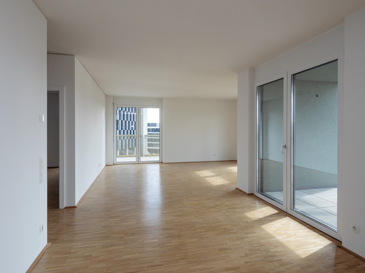 Eine leer stehende Wohnung in einem mehrstöckigen Gebäude. Die Wohnung verfügt über weiße Wände, hellbraune Bodenbeläge und große Glastüren, die zu einem Balkon führen.