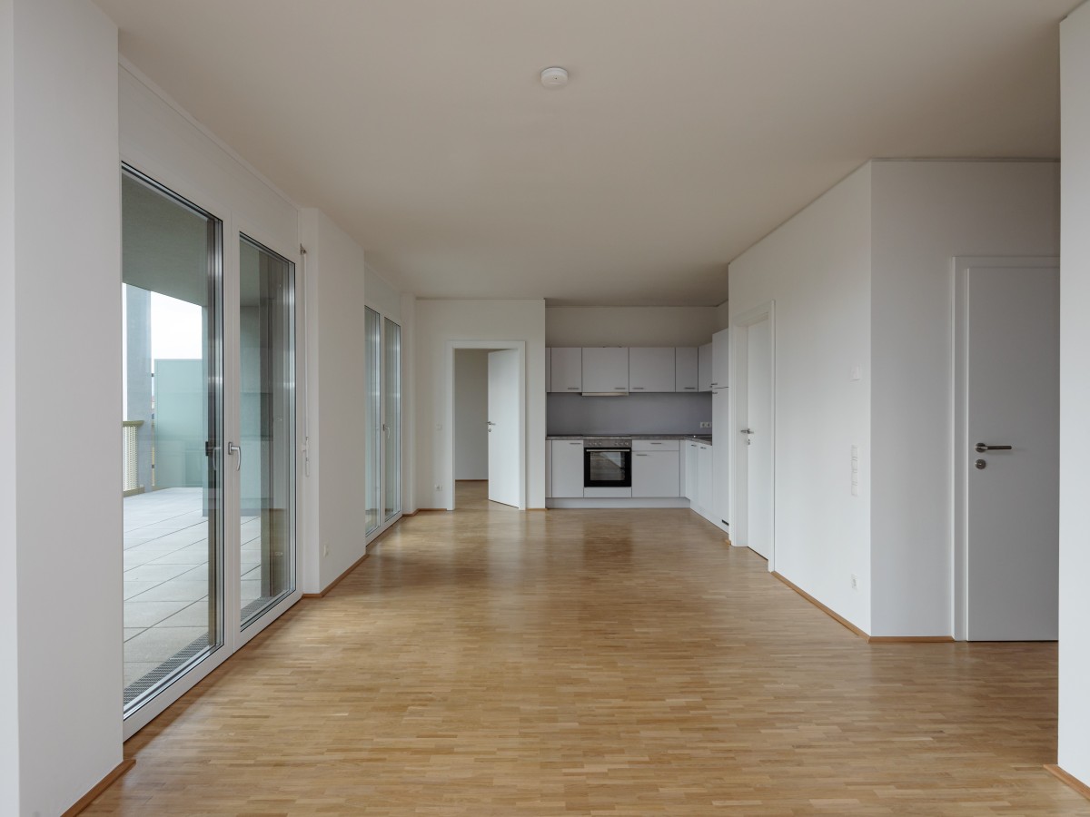 Eine leer stehende Wohnung in einem mehrstöckigen Gebäude. Der offene Wohn- und Küchenbereich hat weiße Wände, hellbraune Bodenbeläge und große Glastüren, die zum Balkon führen.