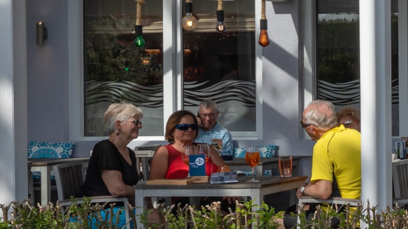 Außenansicht eines Restaurants mit weißer Terrasse, die teilweise überdacht ist und Gästen, die draußen an Tischen sitzen.
