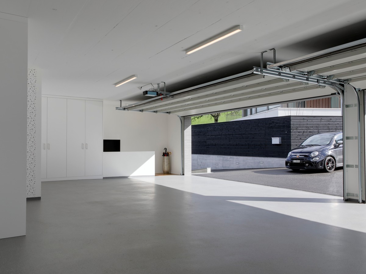 Moderne, offene Garage mit weißen Einbauschränken, grauem Boden und einem Auto vor dem Gebäude.