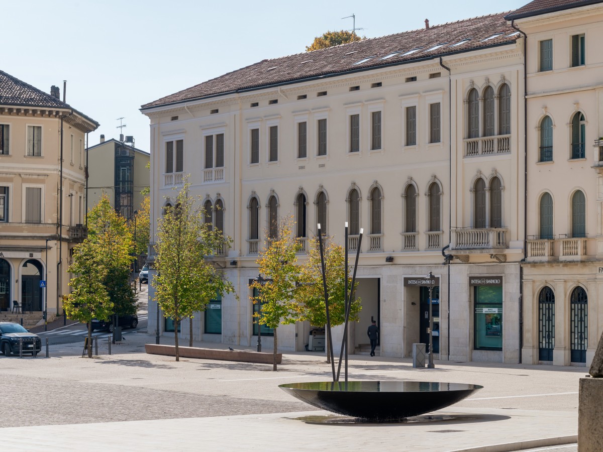  Ein Platz mit einem modernen Brunnen, umgeben von historischen Gebäuden und Bäumen.