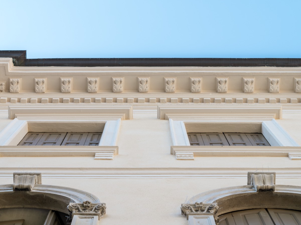 Nahaufnahme eines historischen Gebäudes, das die verzierten Details des oberen Teils und die Fenster mit geschlossenen Fensterläden zeigt.