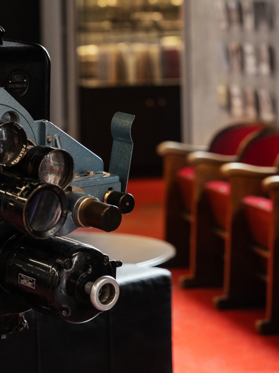 Nahaufnahme eines alten Filmprojektors, der in einem mit rotem Teppich ausgelegten Kinosaal mit roten Kinosesseln steht.