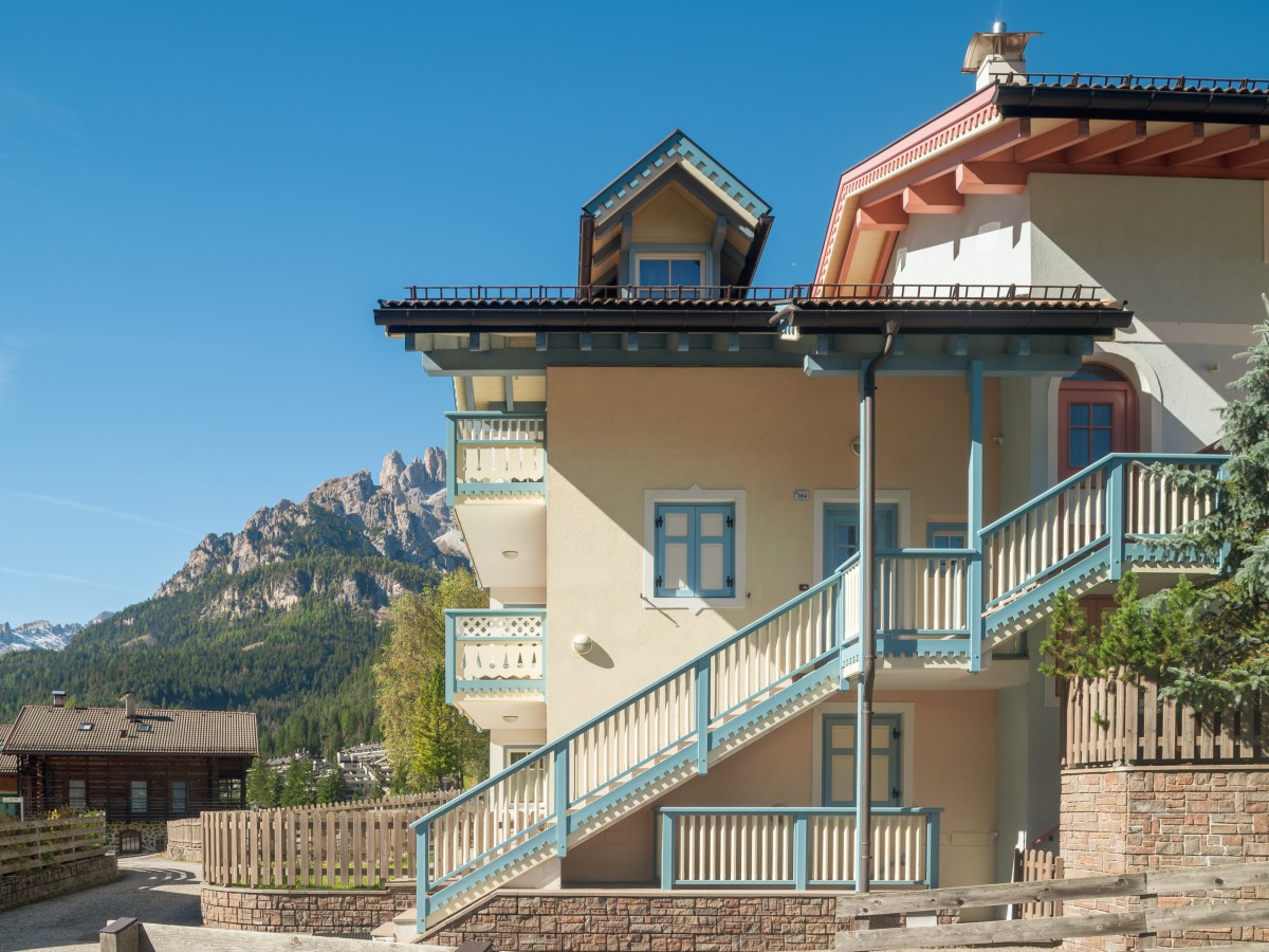 Ein traditionelles Haus im Alpenstil mit einer Außentreppe, mit Bergen und Bäumen im Hintergrund.