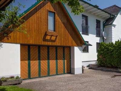  Ein weißes Haus mit einem holzverkleideten Garage und dunkelgrünen Akzenten, umgeben von Bäumen und Sträuchern unter blauem Himmel.