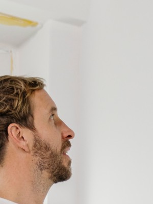 Ein Mann in Arbeitskleidung streicht eine weiße Wand mit einer Malerrolle. Vor ihm stehen zwei Eimer der Marke Brillux, einer davon mit der Aufschrift "Brillux Superlux 3000".