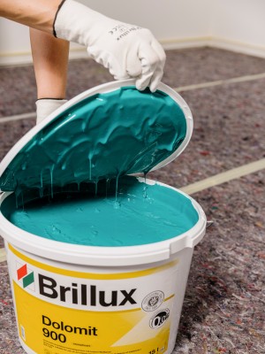  Eine Person in Arbeitshandschuhen hält den geöffneten Deckel eines Brillux-Eimers, aus dem eine leuchtend türkise Farbe tropft. Auf dem Eimer steht „Brillux Dolomit 900“.