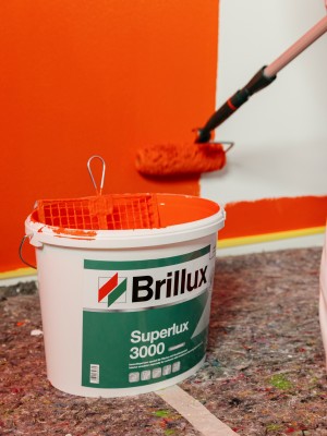 Ein offener Brillux-Eimer mit orangener Farbe und einer Farbrolle davor, neben einer orange gestrichenen Wand.