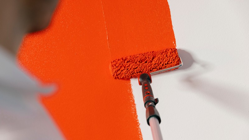  Eine Person streicht mit einer Farbrolle einen breiten Streifen in kräftigem Rot auf eine weiße Wand.