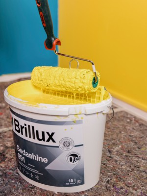 Ein Brillux-Eimer mit gelber Farbe und einer Farbrolle darin, vor einer gelb und blau gestrichenen Wand.