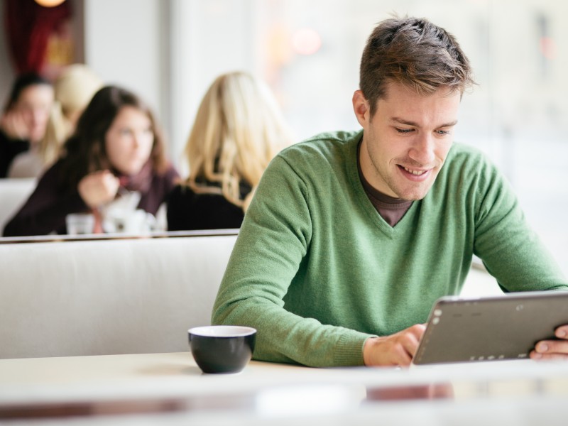 Mann mittleren Alters sitzt in einem Café und nutzt ein iPad, Hintergrund und andere Gäste unscharf